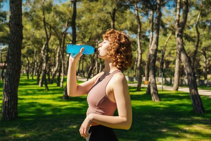 A woman drinking water from a blue bottle in a sunny park.