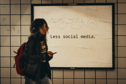 A teenage girl holding a phone in front of a board with the phrase "Less social media".