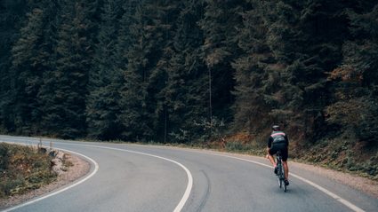 A man cycling on an empty road next to a forest.